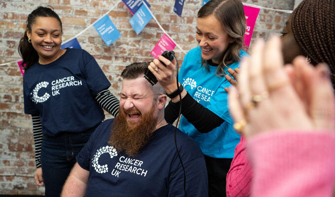 Man getting head shaved during a Cancer Research UK fundraising event, surrounded by smiling supporters